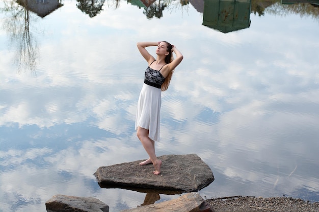 Uma garota de vestido em uma pedra no rio com reflexo do céu com nuvens na água