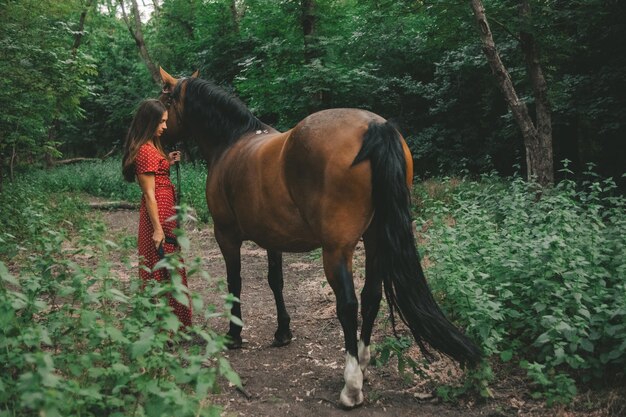 uma garota de vestido vermelho leva um cavalo para passear em um bosque