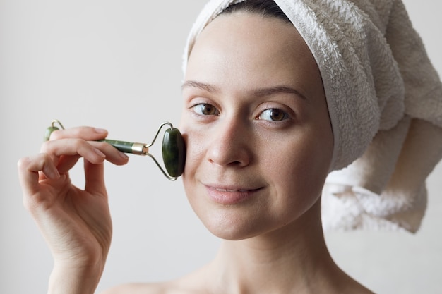 Foto uma garota depois do banho com uma toalha na cabeça faz uma massagem facial com um massagista de jade. foto de alta qualidade