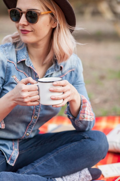 Uma garota desfrutando de uma xícara de bebida quente usando óculos escuros e uma camisa jeans em um piquenique na natureza