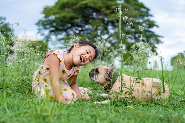 Uma garota e um bulldog francês estão sentados na grama e rindo.