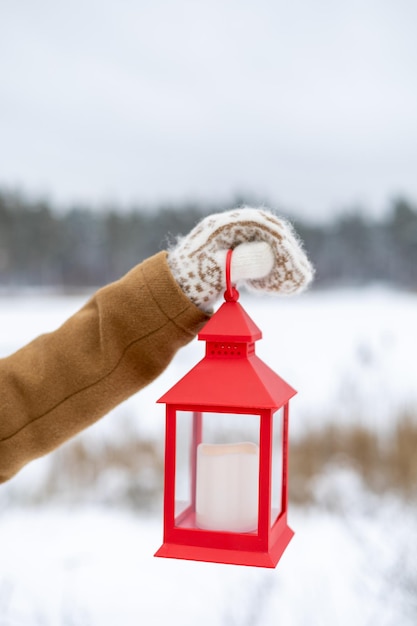 Uma garota em luvas de lã segura uma lanterna vermelha nas mãos com uma vela na floresta de inverno