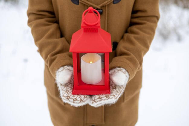 Uma garota em luvas de lã segura uma lanterna vermelha nas mãos com uma vela na floresta de inverno