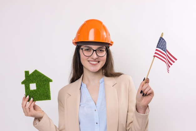 Uma garota em um capacete de construção e uma bandeira americana segura uma casa ecológica verde