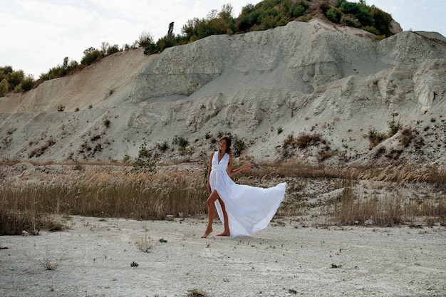 uma garota em um longo vestido branco com flores no cabelo contra o fundo da areia branca da praia e grama seca