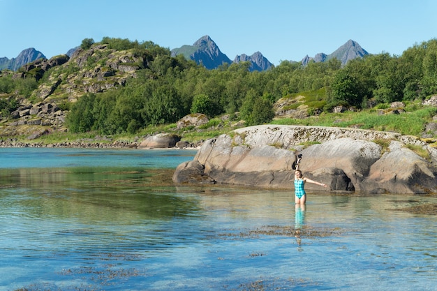 Uma garota em um maiô com uma câmera está na água azul-turquesa transparente da baía, ilha de Arsteinen, arquipélago de Lofoten, Noruega