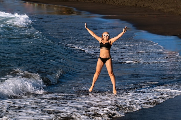 Uma garota em um maiô preto pula na praia.