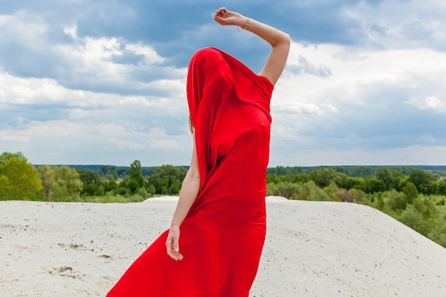 Uma garota em um pano vermelho na areia posa para um fotógrafo no contexto de um céu nublado. Tecido vermelho ao vento abraça a figura da menina.