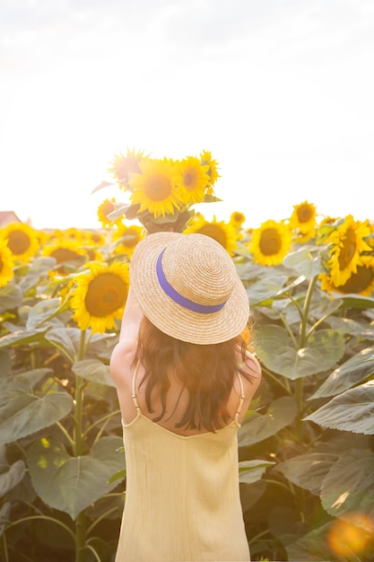 Uma garota em um vestido amarelo e um chapéu de palha fica de costas e segura um buquê de girassóis em um grande campo de girassóis. um lindo dia de sol.
