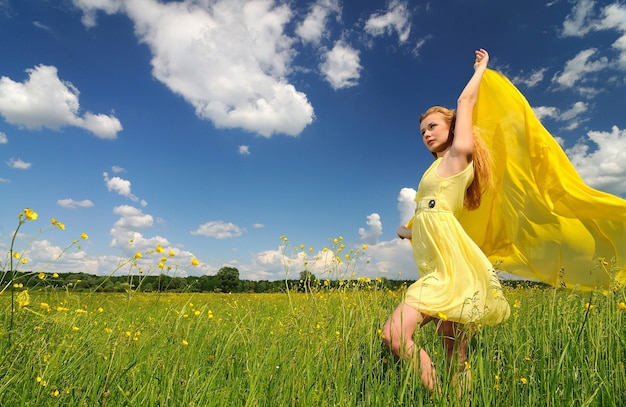 Uma garota em um vestido amarelo posa com as mãos para cima em um campo verde com um pano de seda nas mãos.