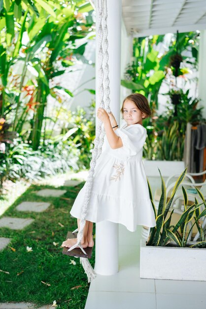 Uma garota em um vestido branco cavalga com os pés em um balanço contra o pano de fundo de um tropical de verão