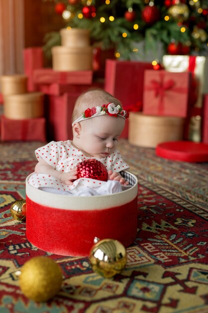 Uma garota em um vestido branco com bolinhas vermelhas está sentada em uma caixa de presente com uma bola de Natal nas mãos