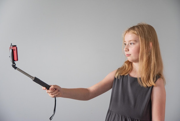 Uma garota em um vestido cinza com um bastão de selfie e um telefone vermelho tira uma foto em um fundo cinza