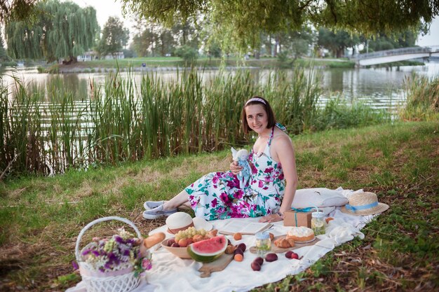 Uma garota em um vestido longo com cabelo curto senta-se em um cobertor branco com frutas e doces, cesta branca com flores.