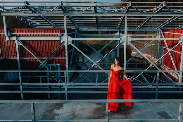 Uma garota em um vestido vermelho em uma barragem perto de um rio ao pôr do sol.