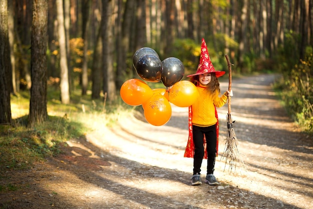 Uma garota em uma fantasia de bruxa e chapéu em uma vassoura com balões laranja e pretos está brincando na floresta de outono indo para uma festa de Halloween