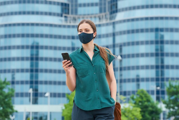 Uma garota em uma máscara facial segurando um smartphone enfiando a mão em um bolso de calças no centro da cidade