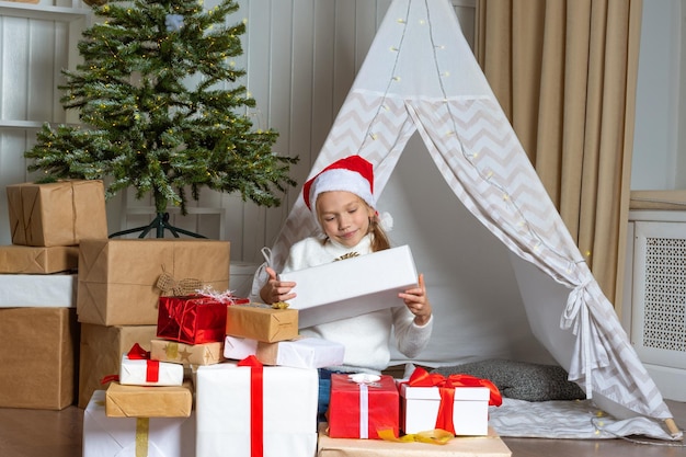 Uma garota emocional com um chapéu de Papai Noel está sentada no chão ao lado de uma pilha de presentes no berçário A criança está feliz com presentes