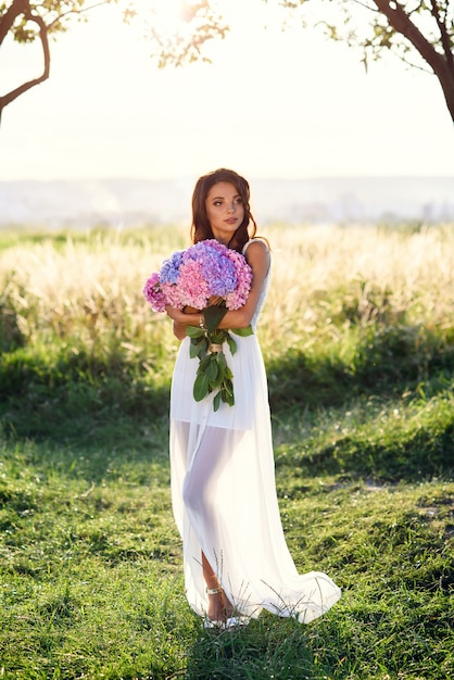 Uma garota encantadora com um sorriso lindo em um vestido branco com um buquê de flores coloridas concurso ao pôr do sol.