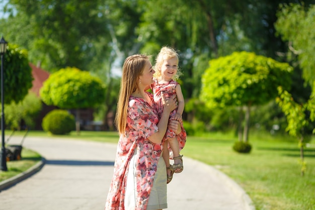 Uma garota encantadora com um vestido de verão leve caminha em um parque verde com sua filha, segurando-a nos braços. Desfruta de dias quentes e ensolarados de verão.