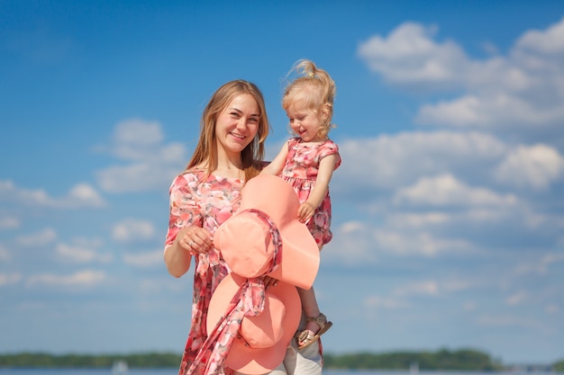 Uma garota encantadora com um vestido de verão leve caminha na praia com sua filha. Desfruta de dias quentes e ensolarados de verão.