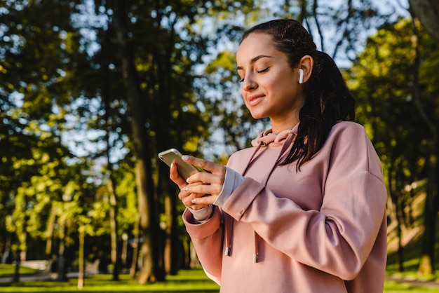 Foto uma garota esportiva sorridente está usando seu telefone com fones de ouvido andando em um parque