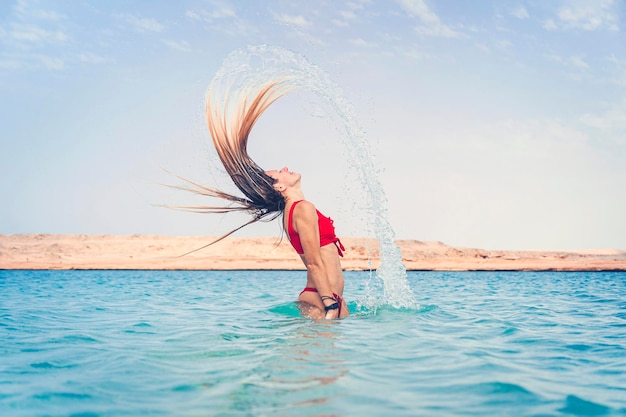Uma garota está se divertindo Uma garota loira em um maiô vermelho espirra na água turquesa no resort contra um céu azul nublado