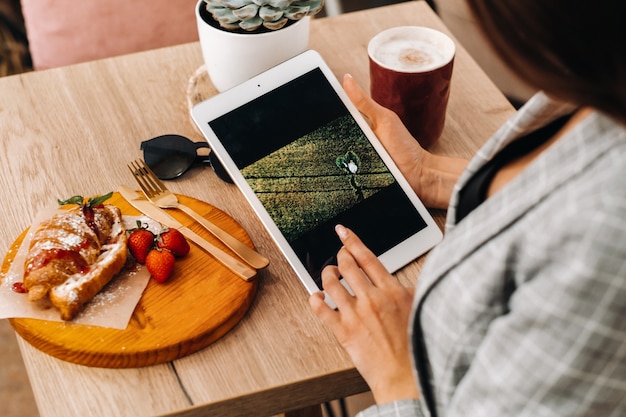 Uma garota está sentada em um café e olhando para um tablet, uma garota em uma cafeteria está sorrindo, Planejando coisas. Há doces na mesa