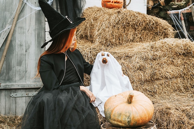 Uma garota fantasiada de bruxa com um cachorro fantasiado de fantasma se divertindo na varanda de uma casa decorada para celebrar uma festa de Halloween