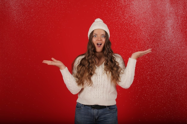 uma garota feliz com um chapéu de inverno e um suéter de tricô em jeans no fundo de neve artificial