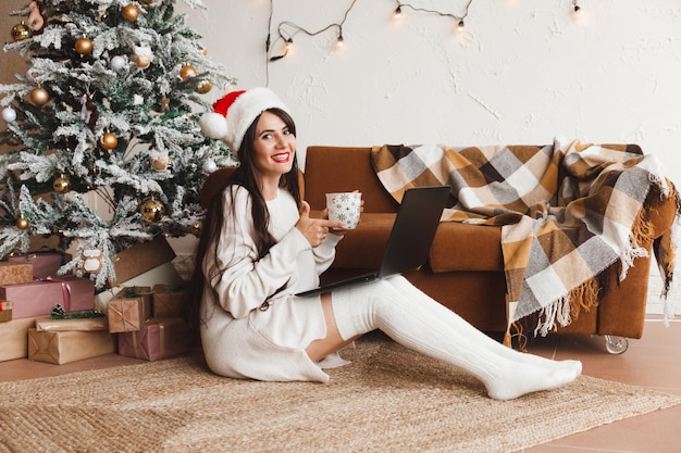 Uma garota feliz com um chapéu de Papai Noel deseja a seus amigos Feliz Natal em um bate-papo por vídeo em um laptop Uma jovem deitada debaixo de uma árvore de Natal entre caixas de presente no interior de uma casa