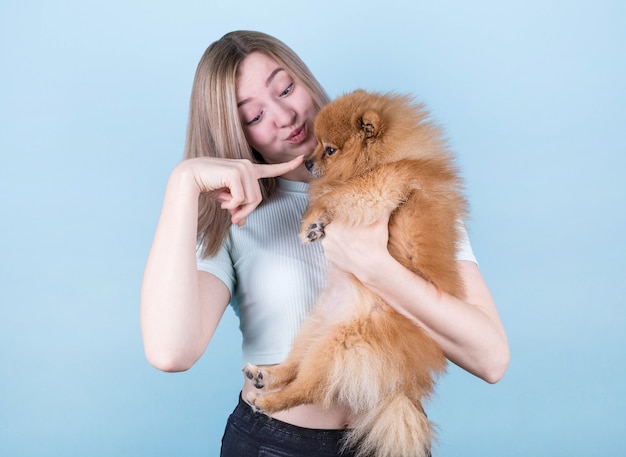 Uma garota feliz e satisfeita recebe um cachorrinho fofo, brinca e abraça carinhosamente seu amigo de quatro patas, fica em pé em uma parede azul, usa uma camiseta curta. uma mulher abraça um pomerânia. pessoas e cachorros