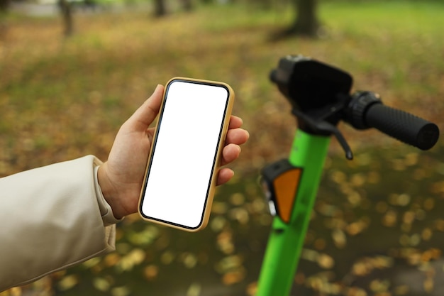 Uma garota fica com uma scooter e mostra um telefone com iluminação branca vazia em um parque de outono