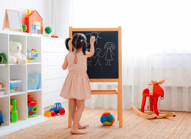 Uma garota fica no quarto das crianças com um quadro-negro e desenha uma família com giz