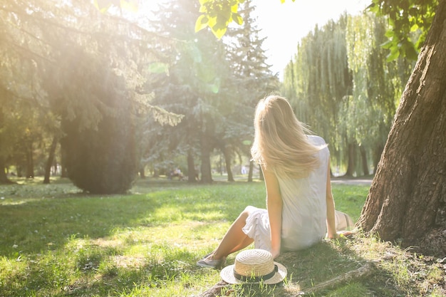 Uma garota hipster elegante com um chapéu de palha está desfrutando do sol ao ar livre Uma mulher feliz sentada em um vestido azul perto da árvore Conceito de férias de viagem Estilo de vida de verão Sombras em árvores Cabelos bonitos