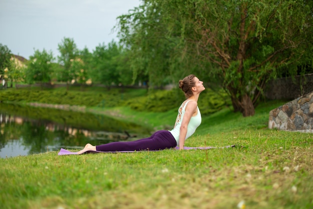 Uma garota jovem esportes pratica ioga em um gramado verde junto ao rio, postura de yoga assans.