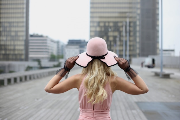 Uma garota loira com cabelos longos em um chapéu rosa e luvas pretas fica para trás