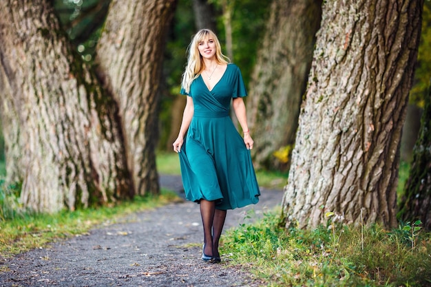 Uma garota loira em um vestido longo verde está andando em um bosque de carvalhos