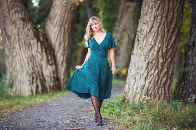 Uma garota loira em um vestido longo verde está andando por um bosque de carvalhos