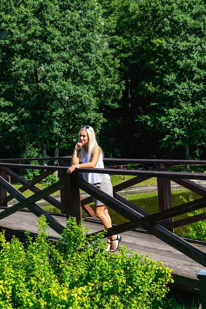 Uma garota loira fica em uma ponte no parque e olha pensativamente para a distânciaFoto vertical