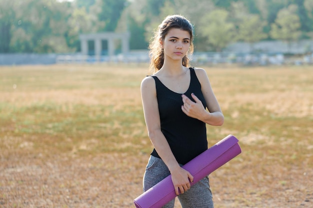 Uma garota magra desportiva em leggings e um top está de pé com um tapete de treinamento, descansando entre os exercícios.
