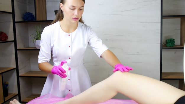 Uma garota, mestra na depilação com açúcar, segura o joelho da modelo com uma das mãos e borrifa um spray sobre ele com a outra para limpá-lo.