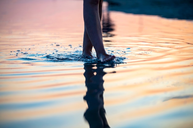 Uma garota na praia ao pôr do sol caminha sobre a água, apenas as pernas são visíveis