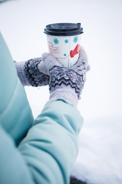 Uma garota no inverno segura uma xícara de café em luvas para aquecer e beber uma bebida quente