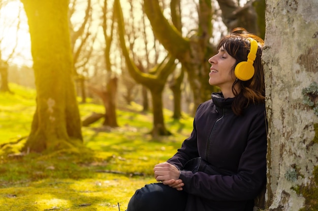 Uma garota ouvindo música com fones de ouvido amarelos na floresta ao pôr do sol sentado em uma árvore com os olhos fechados