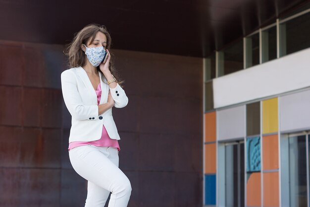 Uma garota real de 40 anos em um terno branco e camisa rosa com cabelo castanho solto e máscara de pano falando no smartphone