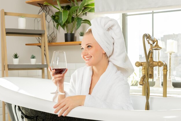 Foto uma garota relaxa no banheiro com uma taça de vinho, uma mulher descansa feliz em um roupão branco