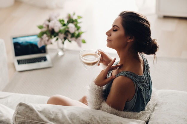 Foto uma garota relaxada em casa bebe café e assiste a um filme. calma doméstica. a garota está sentada confortavelmente no sofá e bebendo café.