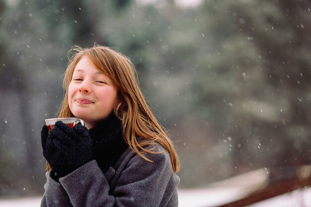 Uma garota ruiva satisfeita com um nariz vermelho em uma floresta de inverno se aquece com um chá quente perfumado