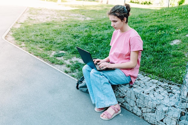 Uma garota se senta em uma pedra em um parque e usa um laptop.